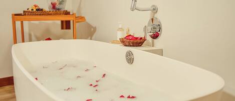 A white freestanding bathtub with rose petals floating in the water, a wooden side table with a bottle of champagne and a basket of fruit, and a small bottle of soap on the wall.