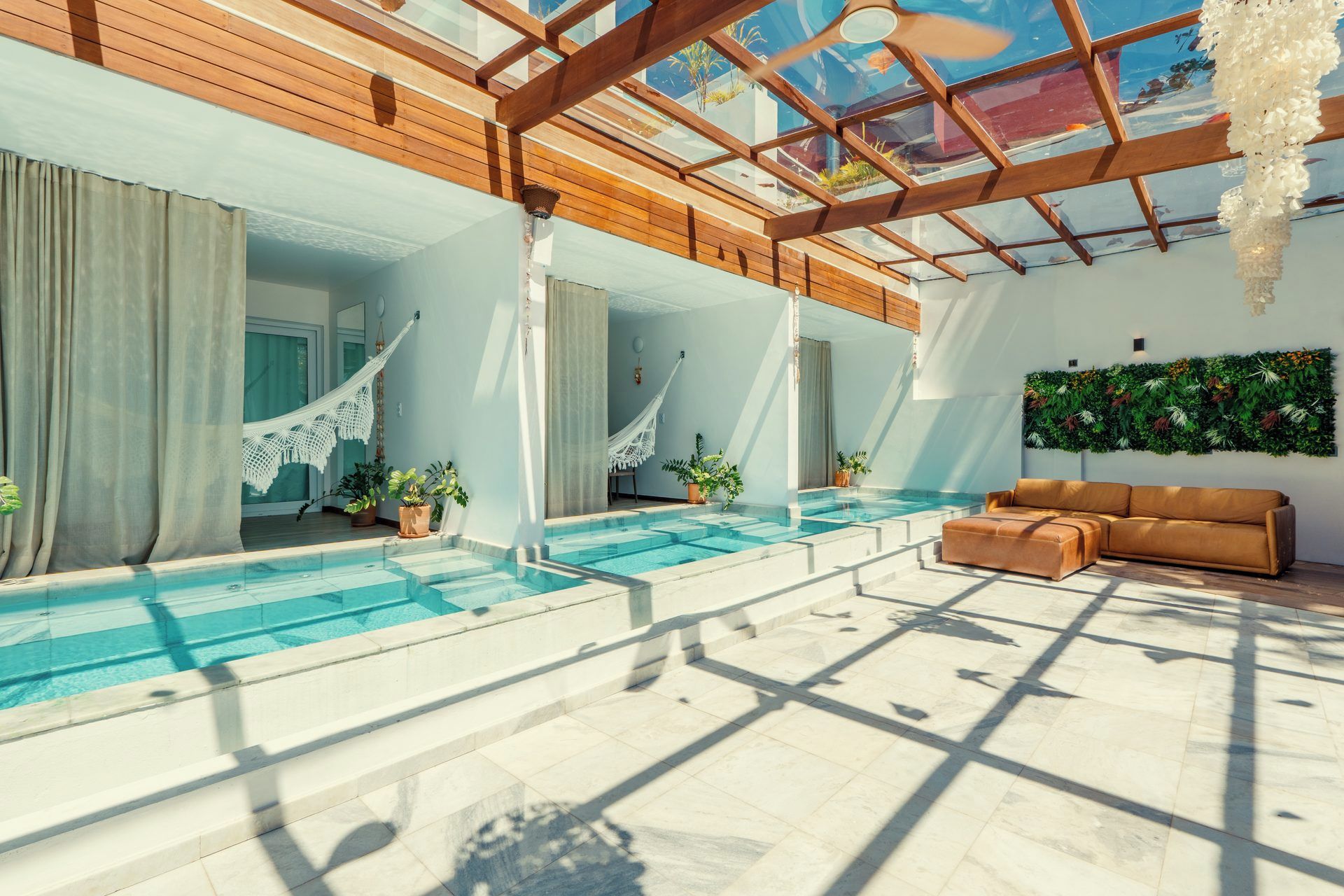 Área moderna de piscina interna com teto de vidro, vigas de madeira e um espaço de estar com sofá.