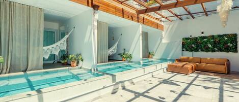 A modern indoor swimming pool area with a glass ceiling, wooden beams, and a seating area with a sofa.