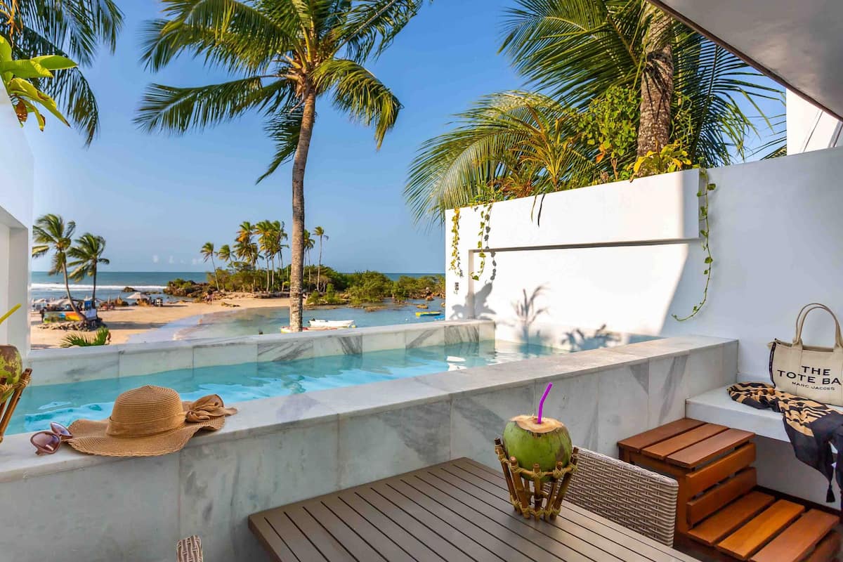 A poolside area with a view of the beach, a lounge chair, a table, and a coconut drink.
