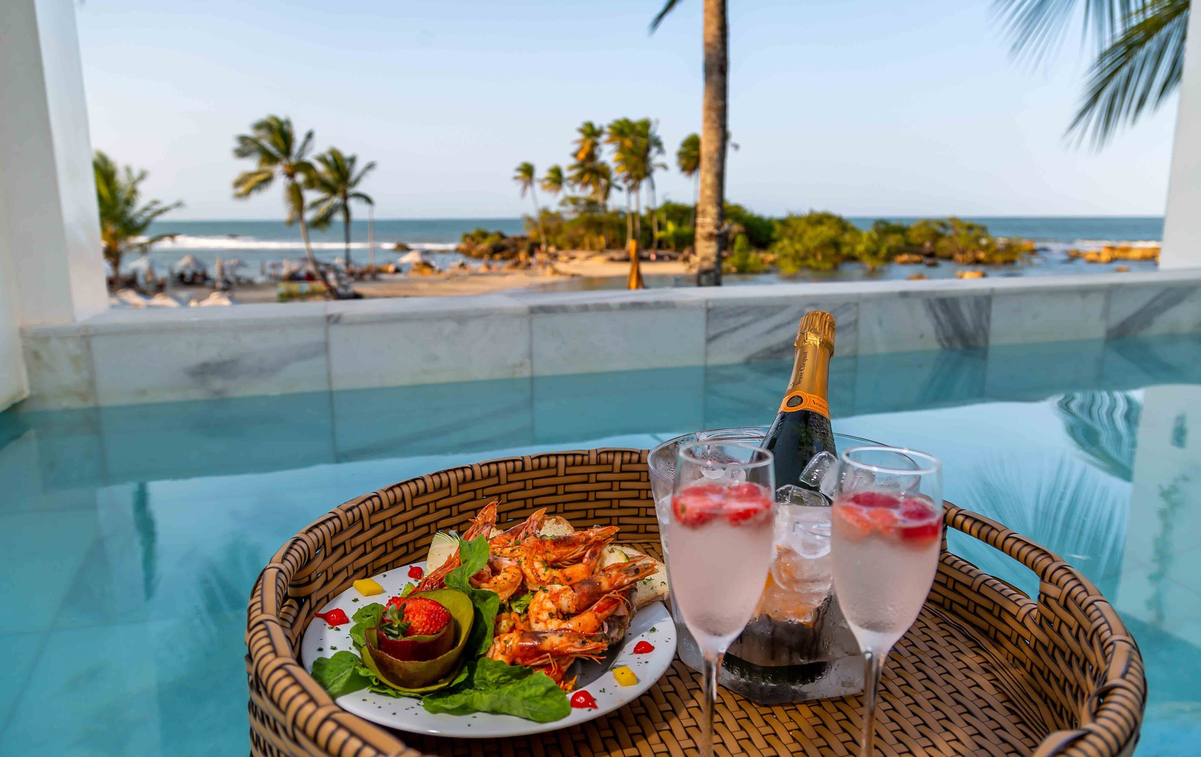 Una mesa de comedor con un plato de camarones, una botella de champán y dos vasos de un cóctel rosado, con vista a una piscina y una playa.