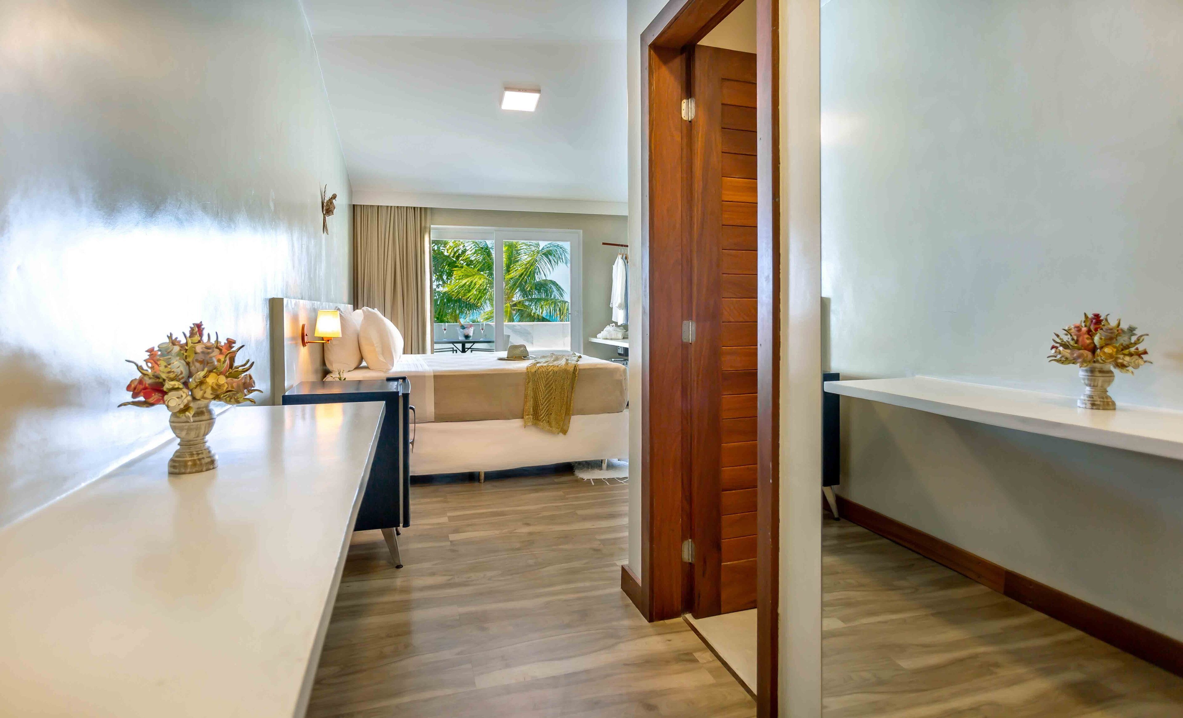 Un hotel moderno con piso de madera, un sofá blanco, un escritorio con un jarrón de flores y vistas a palmeras a través de la ventana.