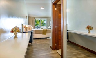 Quarto de hotel moderno com piso de madeira, sofá branco, escrivaninha com um vaso de flores e vista para palmeiras pela janela.