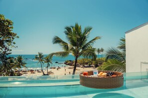 A poolside view with a beach and ocean in the background, a wicker basket with fruits and a drink, and a clear sky. - AQUA BY SAMBASS (Cairu)