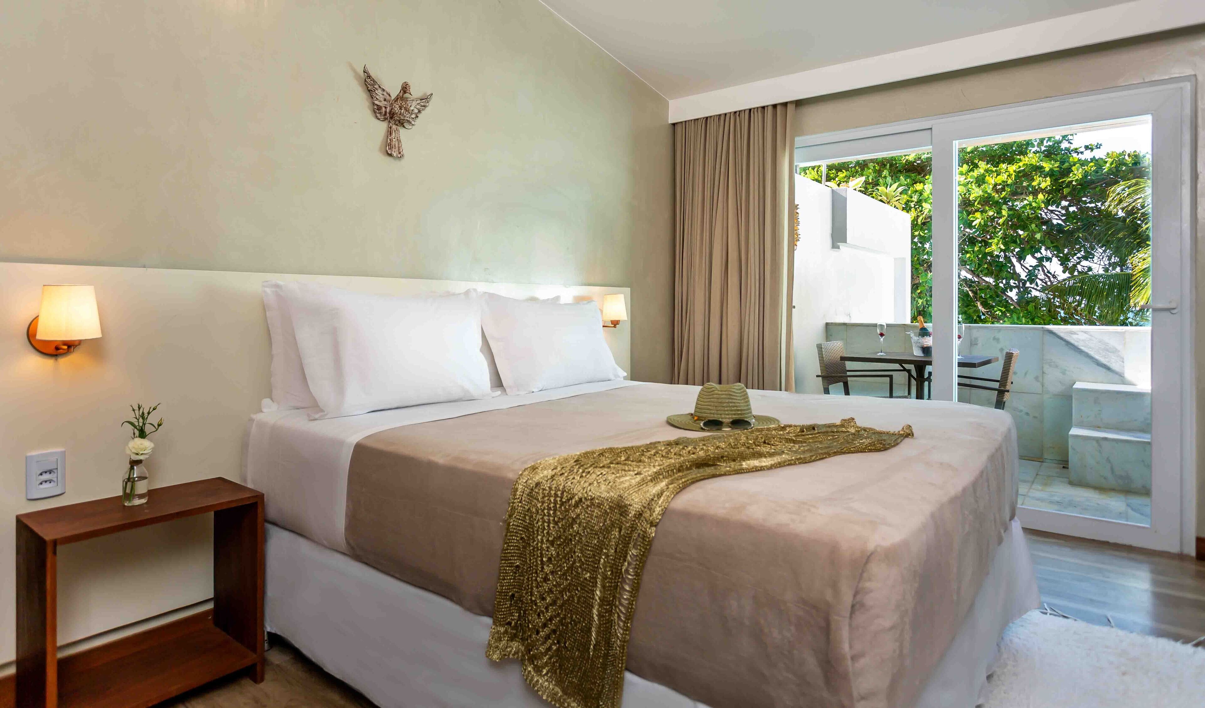 A hotel room with a large bed, a nightstand, a wall-mounted lamp, and a view of the outdoors through a sliding glass door.