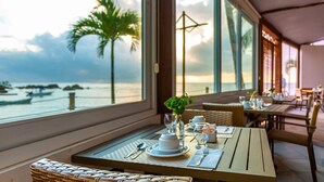 A dining table set with plates, cutlery, and a vase of flowers, overlooking a beach with palm trees and boats. - AQUA BY SAMBASS (Cairu)
