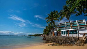 A beachfront hotel with a stone wall, palm trees, and a view of the ocean. - AQUA BY SAMBASS (Cairu)
