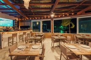 An outdoor dining area with wooden tables and chairs, a bar with a TV, and large windows overlooking a tropical landscape. - AQUA BY SAMBASS (Cairu)
