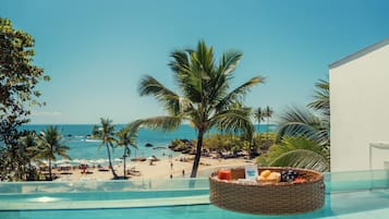 A poolside view with a beach and ocean in the background, a wicker basket with fruits and a drink, and a clear sky.