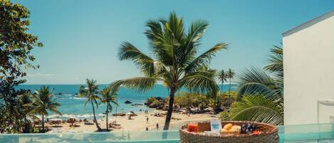 A poolside view with a beach and ocean in the background, a wicker basket with fruits and a drink, and a clear sky.