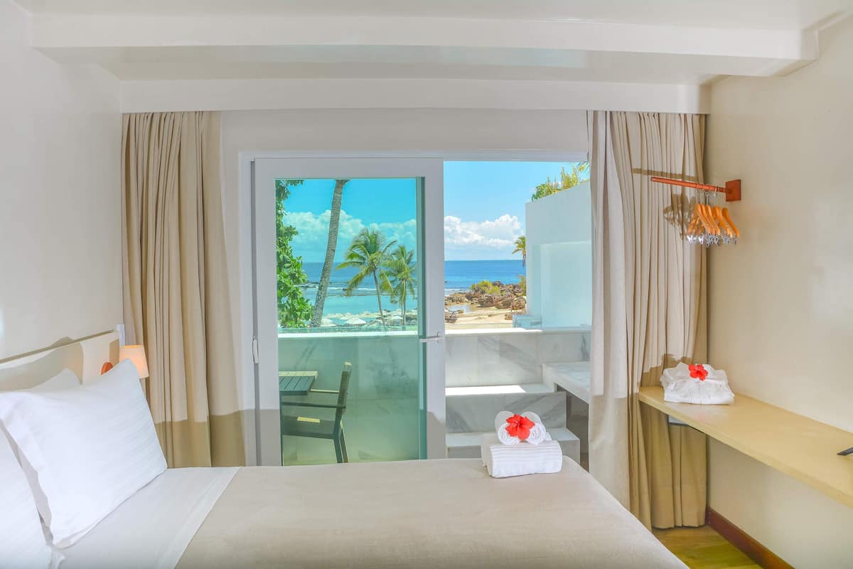 A hotel room with a bed, a nightstand, a lamp, a view of the beach, and a balcony with a table and chairs.