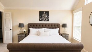 Un letto grande e imbottito con lenzuola bianche, due comodini con lampade, un lampadario e una porta.