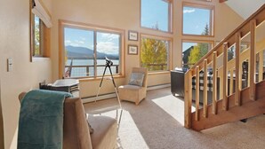 A spacious room with large windows offering a view of a lake and mountains. There is a wooden staircase, a chair, and a tripod.