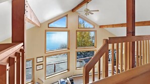 A spacious loft bedroom with large windows, a wooden staircase, and a ceiling fan.