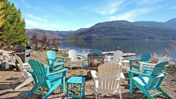 A lakeside seating area with Adirondack chairs and a fire pit, overlooking a calm lake and mountains.