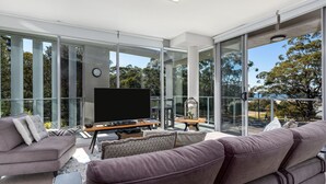 A modern living room with a large flat-screen TV, comfortable seating, and large glass windows offering a view of trees and a body of water. - Husqi Loft - by Latitiude South Coast (Huskisson)