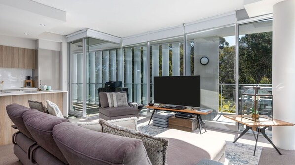 Una sala de estar moderna con sofá, televisión y paredes de vidrio que ofrecen vistas al exterior.