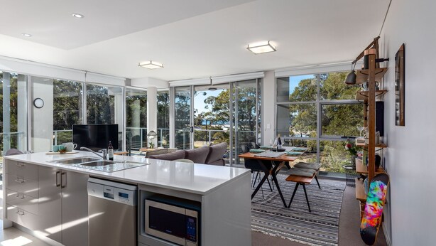 Una cocina moderna con lavavajillas, un comedor con mesa y sillas, y un amplio ventanal con vistas a árboles y una masa de agua.