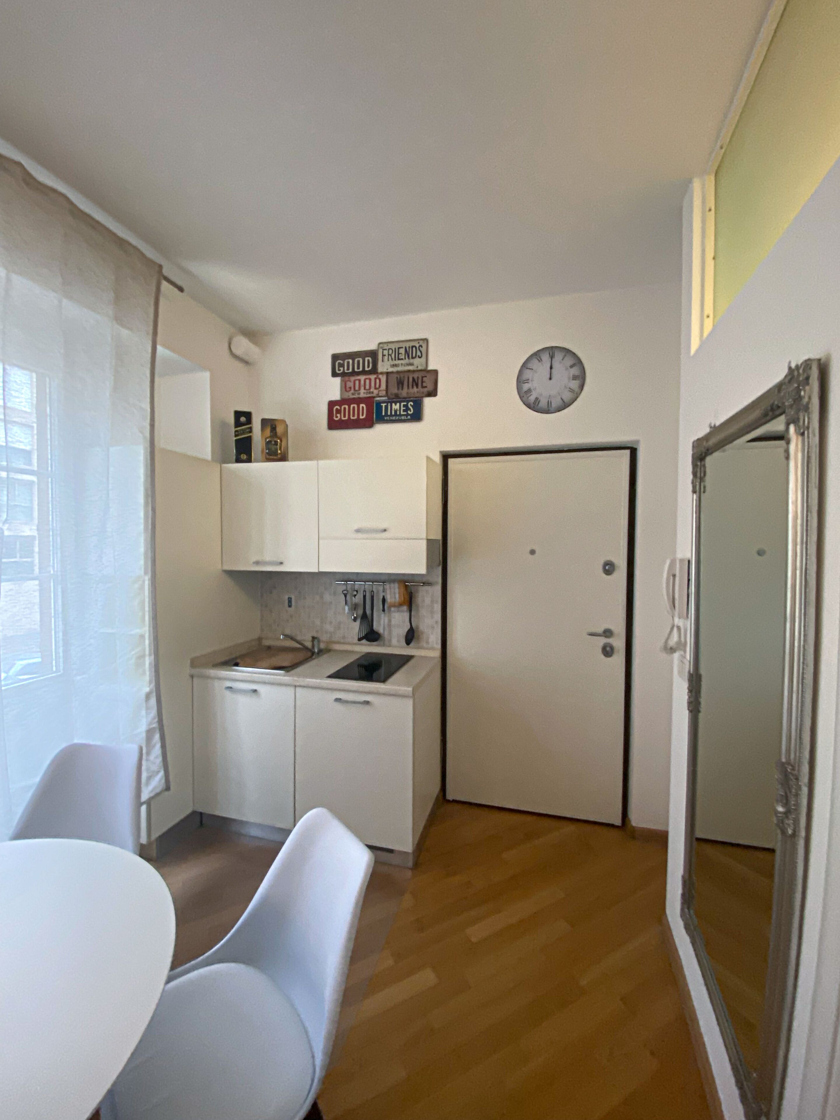 A compact kitchen with white cabinets, a sink, and a clock on the wall. There are signs with phrases like "GOOD FRIENDS" and "GOOD WINE".