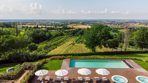Ein Poolbereich mit Liegestühlen und Sonnenschirmen, umgeben von üppiger Bepflanzung und Blick auf weit entfernte Felder und Gebäude.