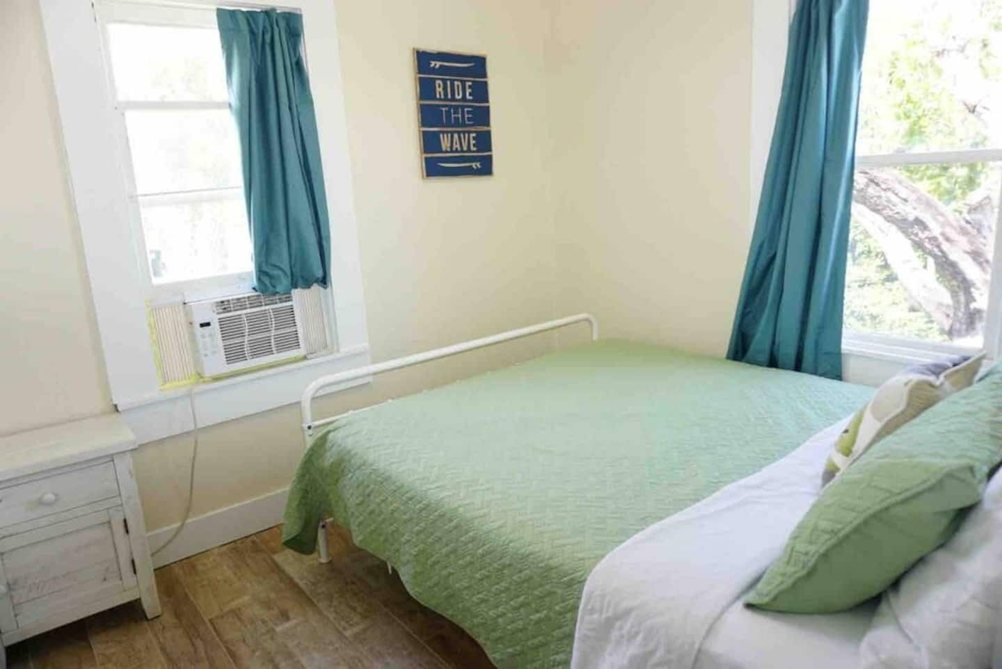 A bedroom with a single bed, a nightstand, a window with curtains, and a wall-mounted air conditioner.