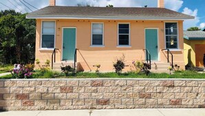Une maison à deux étages avec une façade en pierre, deux portes vertes et un petit jardin devant la maison, agrémenté de plantes.