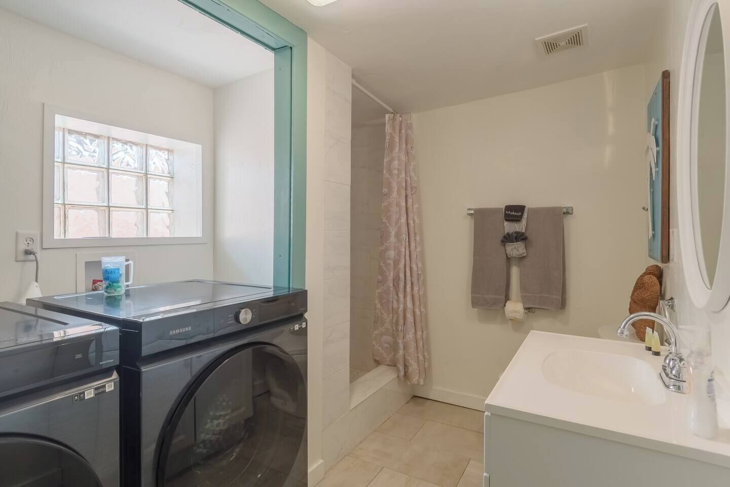A laundry area with a Samsung washing machine, a window with blinds, and a shower with a pink curtain.