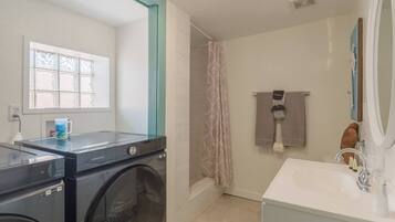 A laundry area with a Samsung washing machine, a window with blinds, and a shower with a pink curtain.