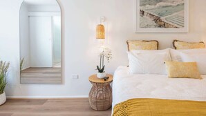 A bedroom with a large bed, a bedside lamp, a framed picture of a coastal scene, and a mirror reflecting the room.
