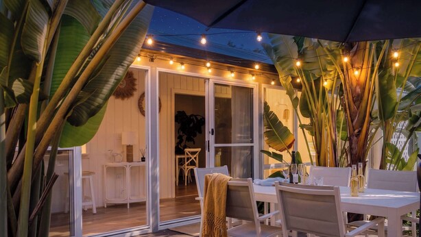 An outdoor dining area with white furniture, string lights, and tropical plants.