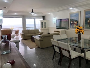 Uma sala de estar espaçosa com um conjunto de sofás, uma mesa de jantar de vidro e uma TV fixada na parede.