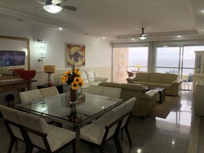 Uma sala de estar moderna com uma mesa de jantar de vidro, sofás brancos e uma grande janela com vista para o mar.