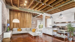 A spacious living area with a wooden ceiling, a white sofa with yellow cushions, a large window with curtains, and a kitchen area with stainless steel appliances. - New York-Style Loft in Sydney CBD with City Views (Darlinghurst)