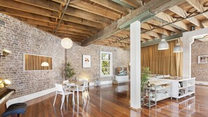 A spacious room with exposed brick walls, wooden beams, a piano, a dining area with white chairs, and a white console table. - New York-Style Loft in Sydney CBD with City Views (Darlinghurst)