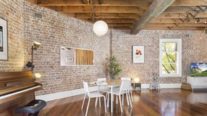 A room with a brick wall, wooden beams, a piano, a dining table with chairs, a plant, and a TV. - New York-Style Loft in Sydney CBD with City Views (Darlinghurst)