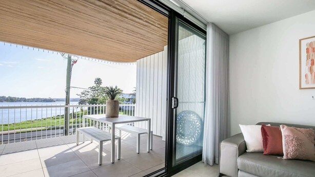 A balcony with a view of a body of water, a white table with a plant, and a grey sofa with cushions.