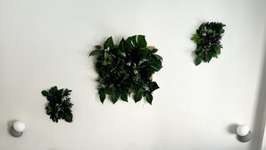Un lit bien fait, avec du linge de lit blanc et deux coussins décoratifs, une plante fixée au mur et une paire de serviettes pliées posées sur un chemin de lit beige.