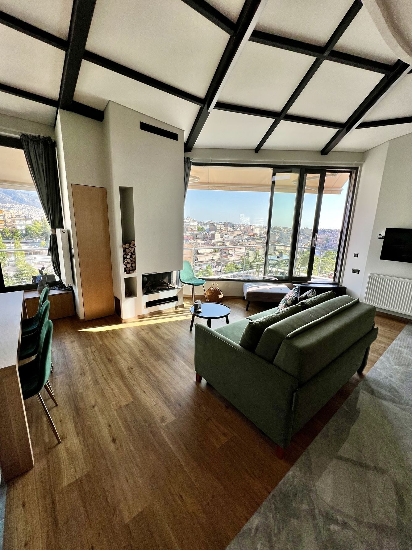 A modern living room with a large sofa, a dining area with chairs, a TV, and a city view through large windows.