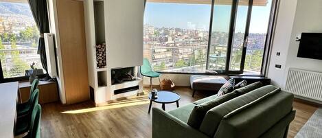 A modern living room with a large sofa, a dining area with chairs, a TV, and a city view through large windows.