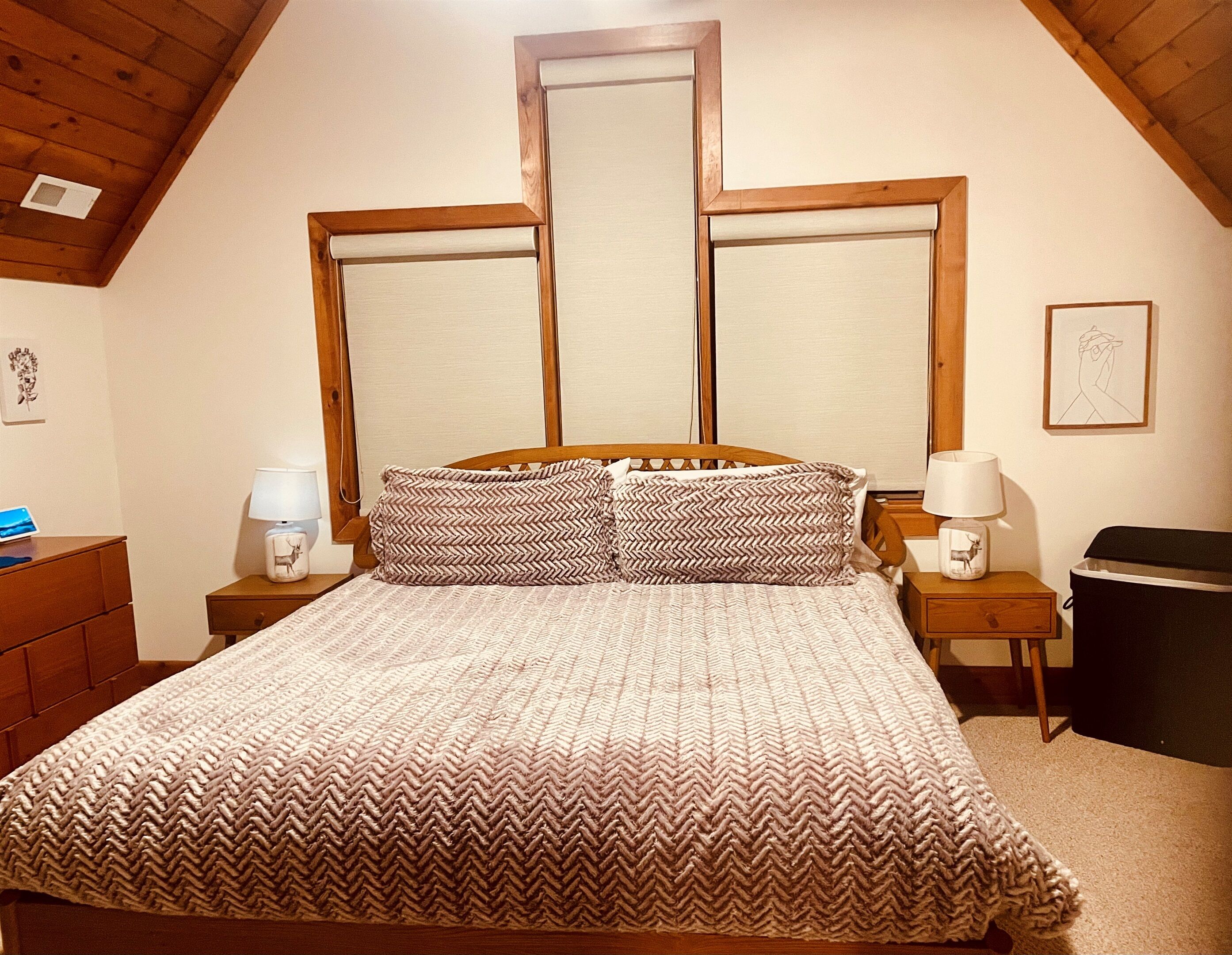A bedroom with a wooden ceiling, a bed with patterned bedding, two bedside tables with lamps, and a framed picture on the wall.