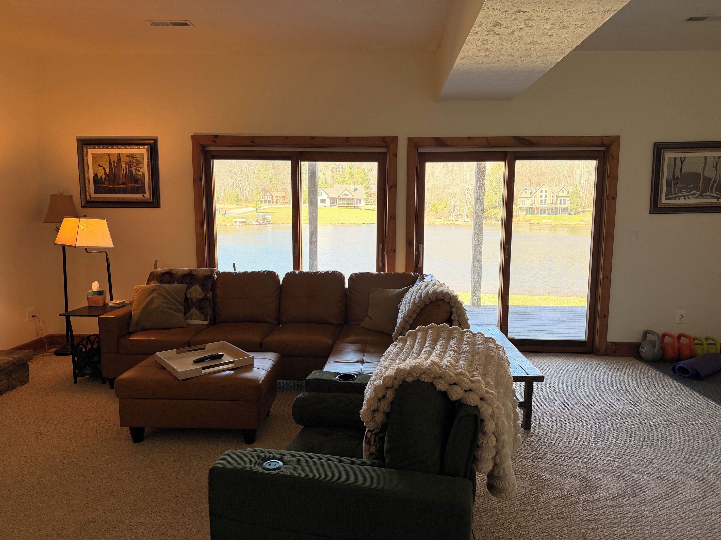 A living room with a sofa, ottoman, and armchair, a lamp, and large windows with a view of a lake and houses.