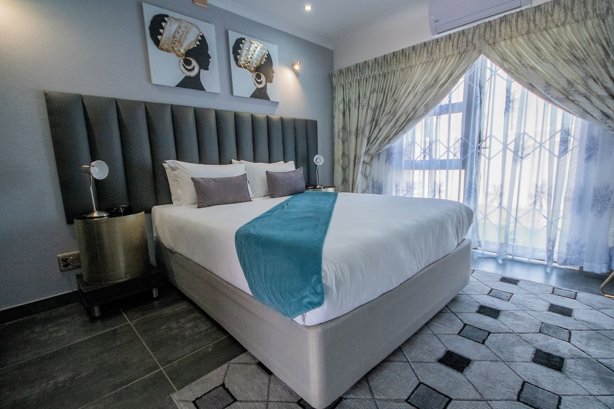 Una camera d'albergo moderna con un letto grande, una testiera, lampade sui comodini e vista all'esterno tramite un'ampia finestra.