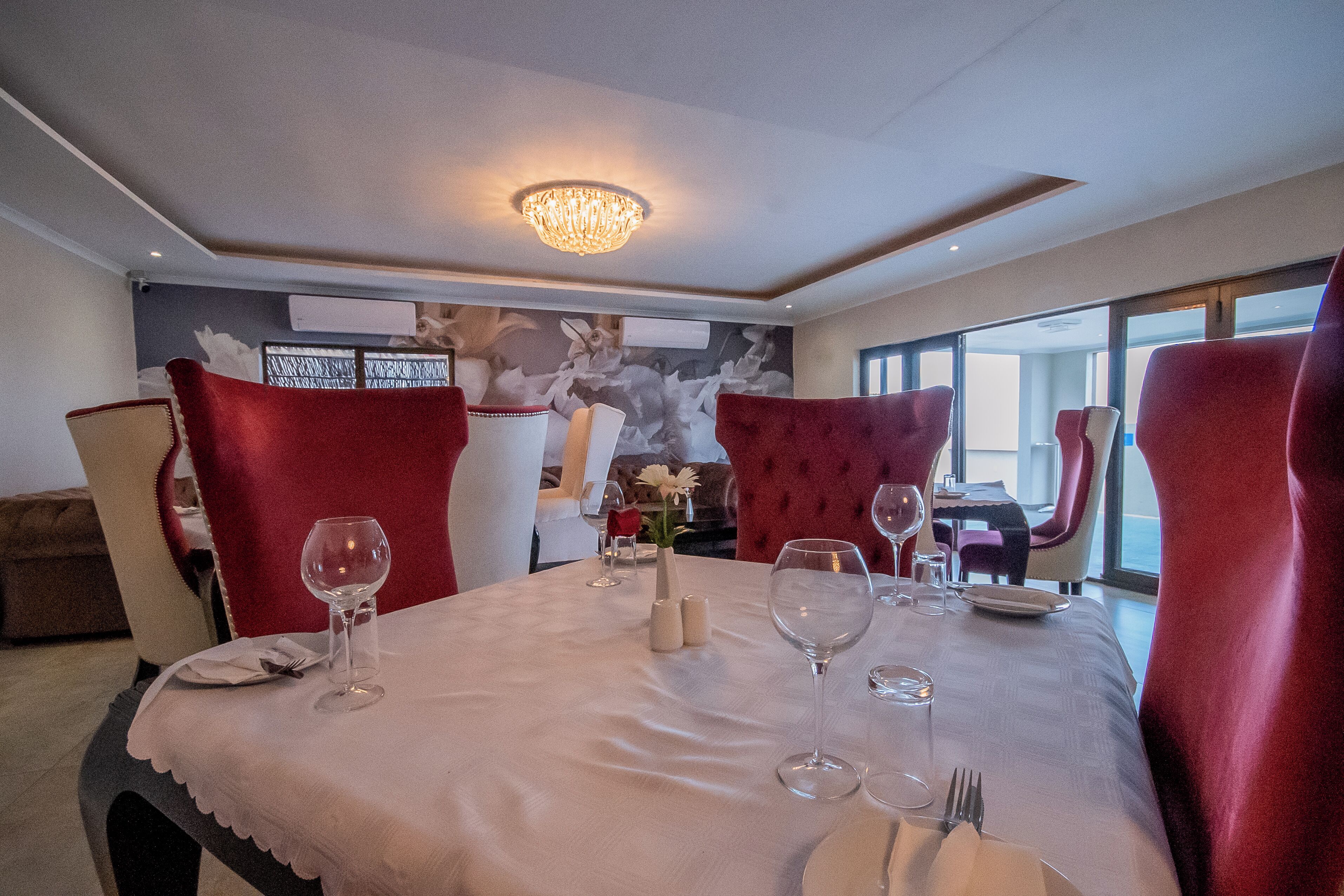 A dining area with a chandelier, red upholstered chairs, a white tablecloth, and a vase with flowers.