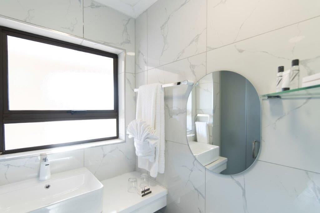 Une salle de bain moderne avec un grand miroir, des carreaux de marbre blanc, un lavabo blanc et une douche en verre.