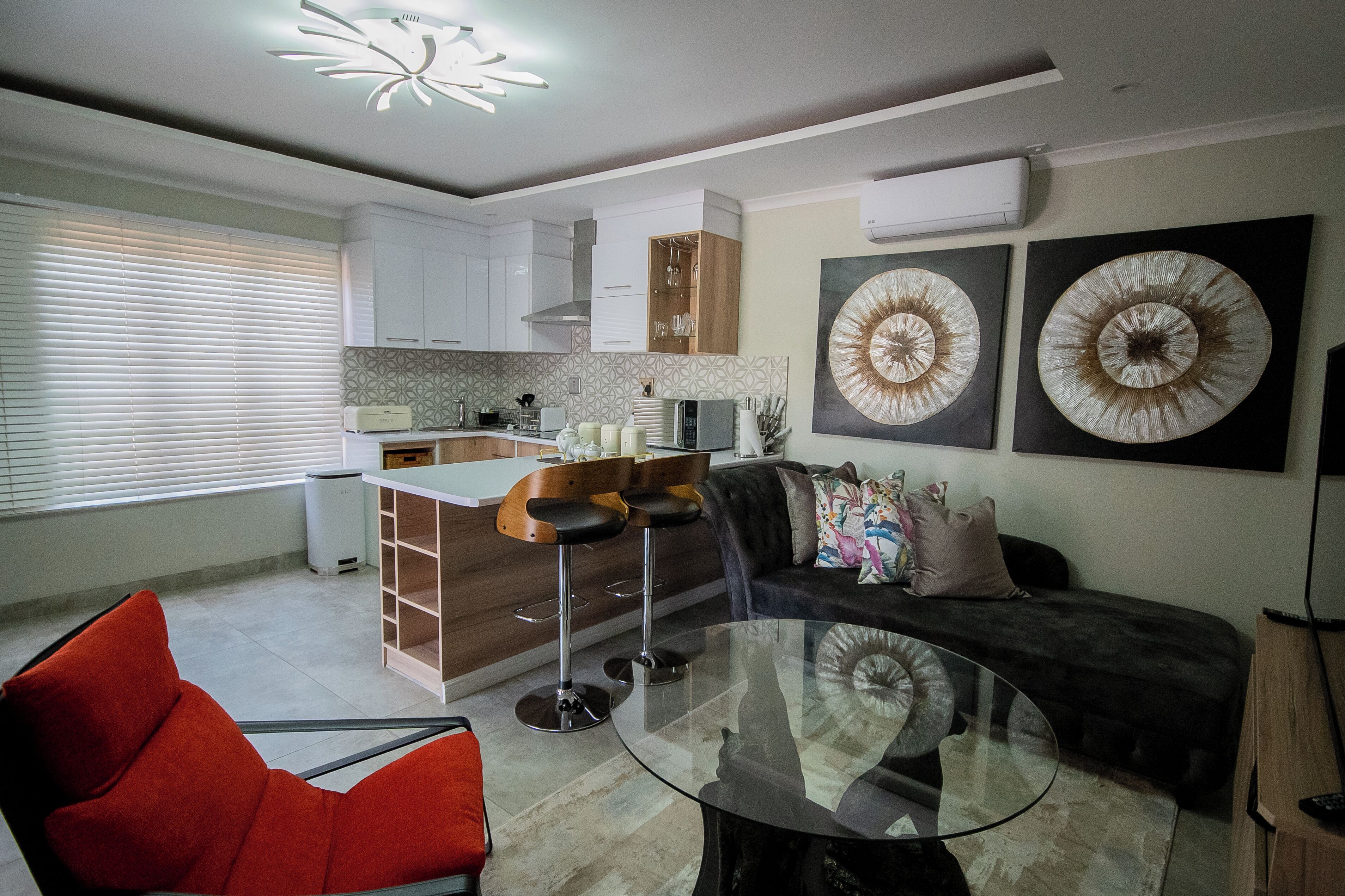 Una sala moderna con una mesa de centro de vidrio, una silla roja, un sofá oscuro y una cocina con armarios blancos y una isla de madera.