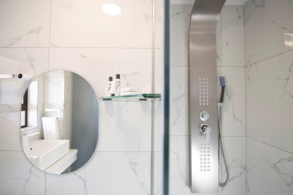 Ein modernes Badezimmer mit einer Glaskabine zur Dusche, einem runden Spiegel und einer Wand mit weißen Marmorfliesen.