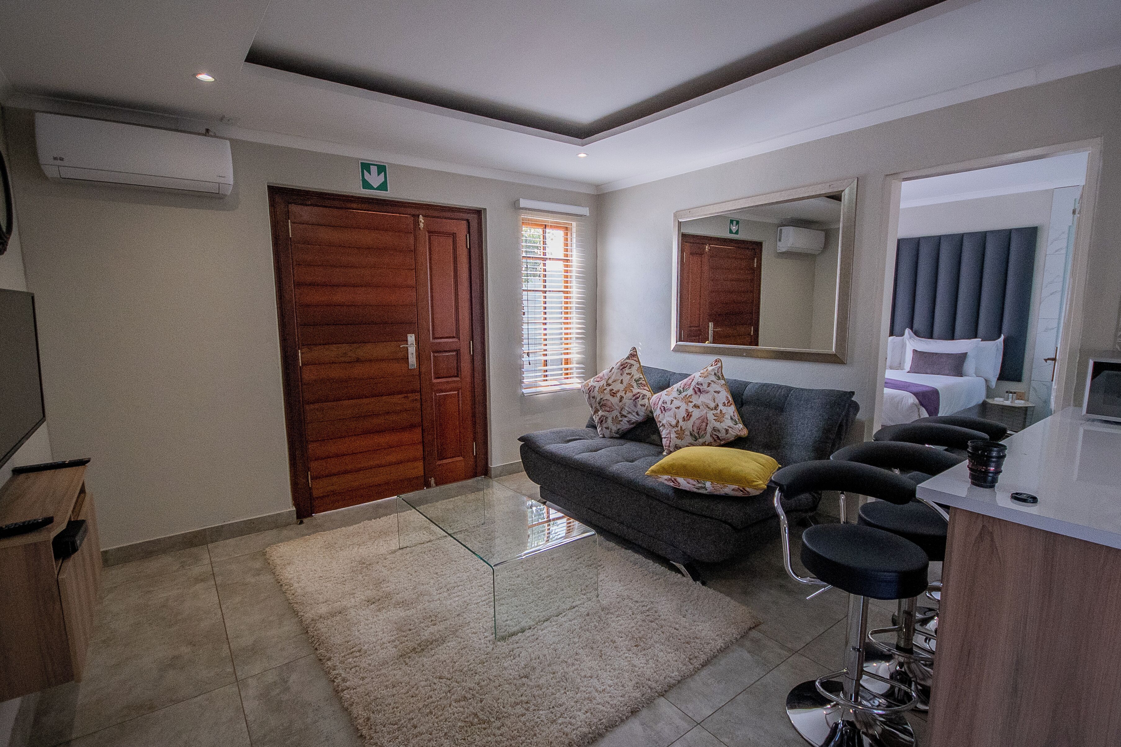 Una camera d'albergo moderna con un divano grigio, un piccolo angolo bar, uno specchio grande e una porta in legno.
