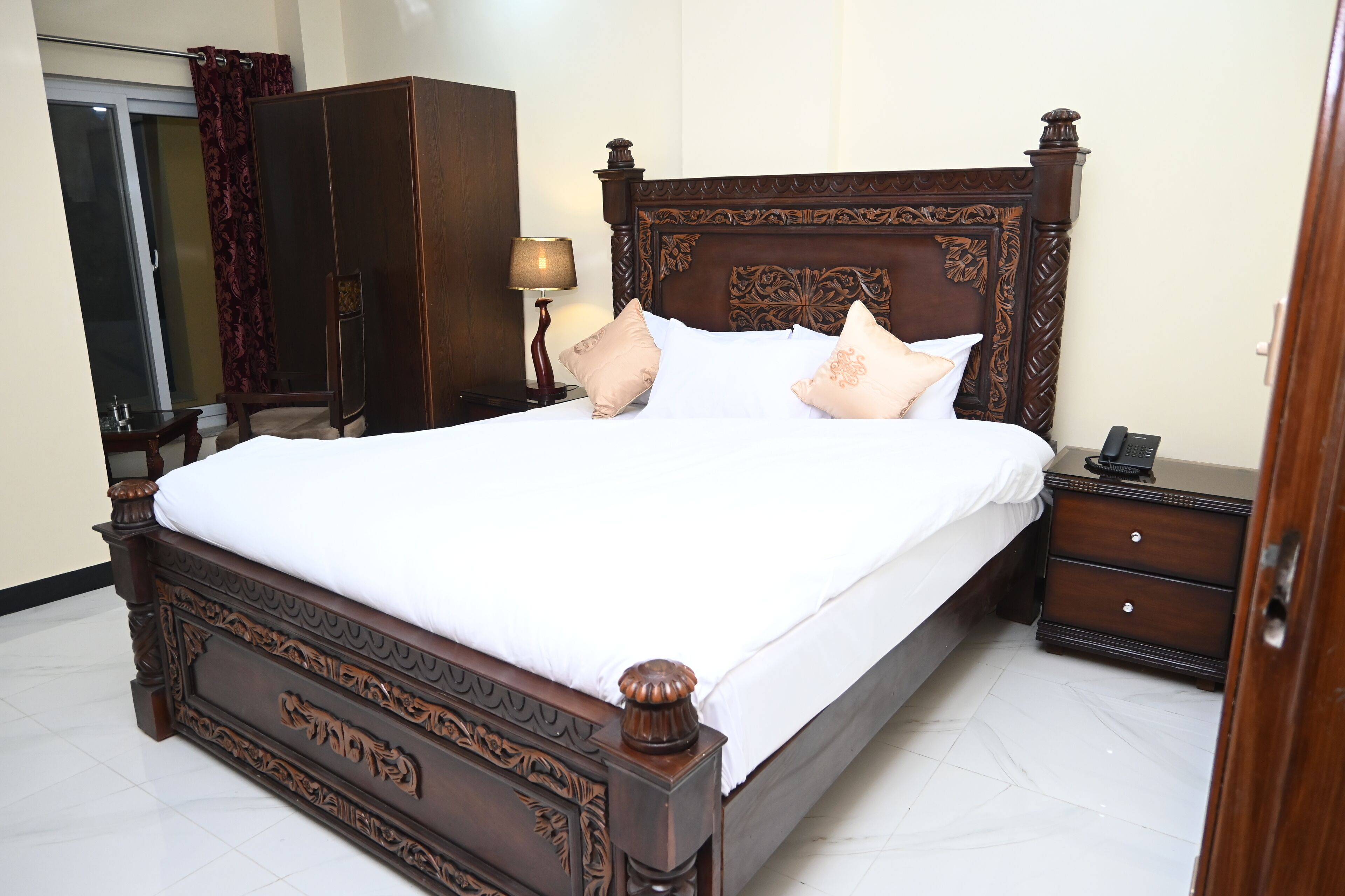 A neatly made bed with a wooden headboard, a nightstand with a lamp, and a wardrobe in a hotel room.