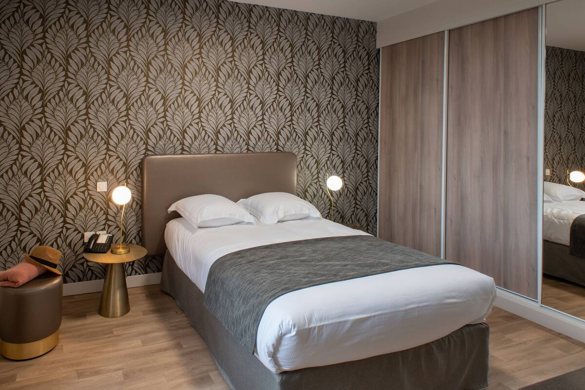 A modern bedroom with a large bed, a bedside table with a lamp, a mirror, and a patterned wallpaper.