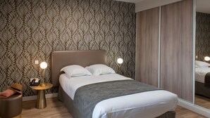 A neatly made bed with a grey blanket, a bedside lamp, and a telephone on a small round table beside it.